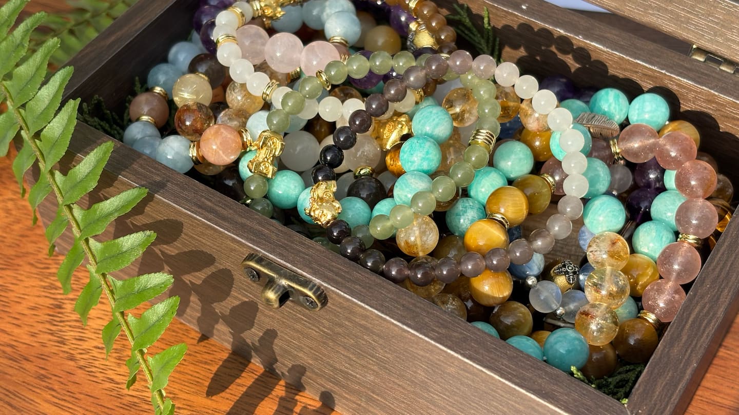 Stack of colourful beaded bracelets in a wooden box with a leafy green plant on the side.